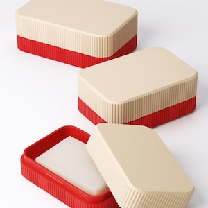 Può includere: Tre portasapone rettangolari con design bicolore. La parte superiore e il coperchio sono beige, mentre la base è rossa. Un coperchio è aperto, rivelando una saponetta bianca. I portasapone hanno una texture a coste.