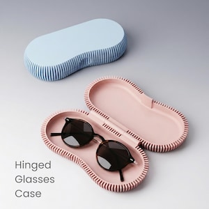 Puede incluir: Un estuche de gafas rosa claro con gafas de sol redondas en su interior. El estuche está abierto, revelando el interior y las gafas. La tapa es azul claro con rayas azules verticales. El texto "Hinged Glasses Case" es visible.