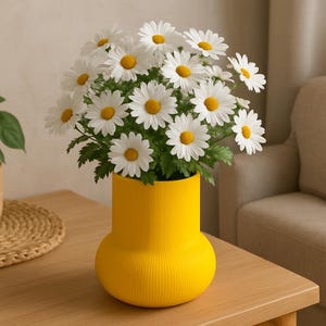 May include: A bright yellow vase with a ribbed design contains a collection of white daisies with yellow centres and green leaves. The vase is placed on a light wooden table, creating a cheerful display.