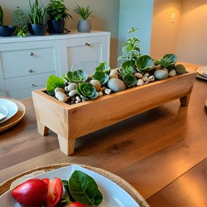 May include: A rectangular wooden planter box filled with green succulents, small stones, and soil. The planter sits on four short legs and is placed on a wooden table. The table is set with plates and silverware.