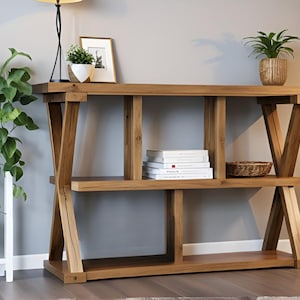 Op de afbeelding: Een houten console met een natuurlijke afwerking. De tafel heeft een ontwerp met twee niveaus met een open plank en een bovenblad. Decoratieve items zijn een ingelijste foto, potplanten en boeken. De poten van de tafel hebben een X-vormig ontwerp.
