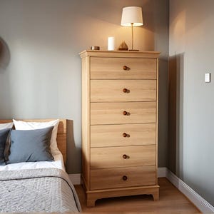 Puede incluir: Una cómoda alta de madera clara con seis cajones y pomos redondos y oscuros. La cómoda está en un dormitorio, junto a una cama con almohadas azules y un edredón gris. Una lámpara y velas están encima de la cómoda.