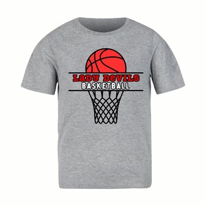 Op de afbeelding: Grijs t-shirt met een rood basketbalontwerp boven een zwart basketbalnet. De tekst "Lady Devils Basketball" is in het rood gedrukt. Het t-shirt is gemaakt van zacht materiaal.
