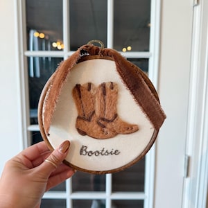 May include: An embroidered hoop art piece with a pair of brown cowboy boots on cream fabric. The word "Bootsie" is stitched below the boots. A brown ribbon is attached to the top of the hoop, with a wooden frame.