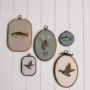 May include: Five wall-mounted embroidery hoops featuring needle-felted wildlife designs. The collection includes a trout on beige fabric, a deer head with the text Abel James on sage green, a small trout on blue, a mallard on striped fabric, and a mallard on cream.