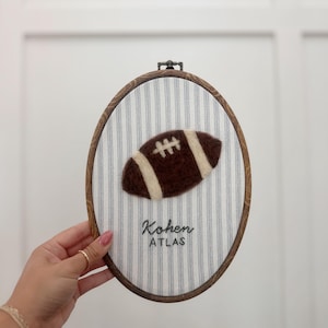 May include: Oval-shaped wall hanging featuring a brown felt football with white stripes, set against a striped fabric background. The wooden frame holds the football and fabric. The name "Kohen ATLAS" is embroidered below the football.