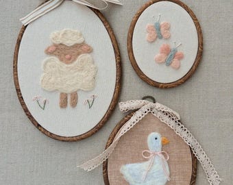 May include: Three oval-shaped wall hangings with embroidered animal designs. One features a sheep, another two butterflies, and the last a goose. Each is framed in wood and adorned with a ribbon or lace bow.