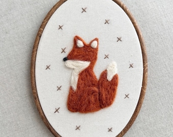 May include: An oval-shaped embroidery hoop featuring a handcrafted fox design. The fox is rendered in shades of orange and white, with a black nose, set against a cream-colored fabric. Brown 'x' stitches surround the fox, and the hoop has a wooden frame.