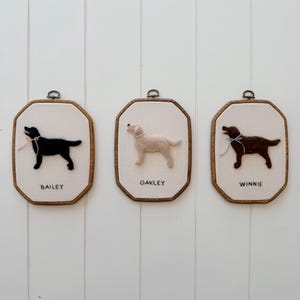 Puede incluir: Tres marcos de madera octogonales que contienen retratos de perros hechos con fieltro de aguja sobre tela blanca. El marco izquierdo muestra un perro negro con un lazo rosa etiquetado BAILEY. El marco central muestra un perro crema etiquetado OAKLEY. El marco derecho muestra un perro marrón con un lazo azul etiquetado WINNIE.