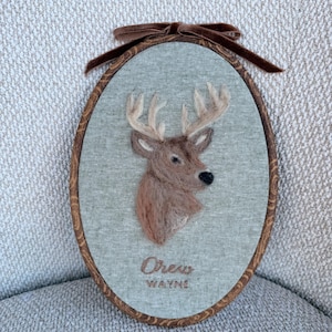 May include: An oval framed artwork featuring a deer head with antlers. The deer is rendered in shades of brown and tan, set against a light green fabric. The frame is wood-toned, and a brown velvet bow adorns the top. The text "Drew Wayne" is embroidered below the deer.