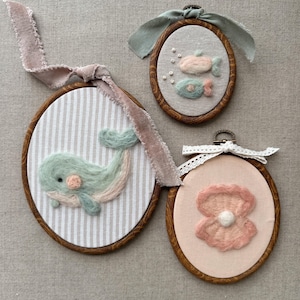 May include: Three oval-shaped wall hangings with embroidered sea-themed designs. One features a whale, another two fish, and the last a pearl. Each is framed in wood and has a ribbon for hanging. The whale is light blue and pink.
