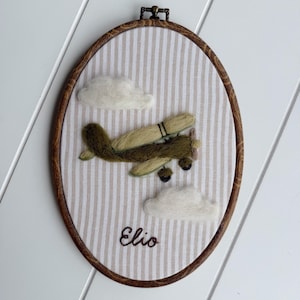 Puede incluir: Un bastidor de bordado ovalado con tela de rayas verticales beige y blancas. Un avi&oacute;n verde oliva de lana fieltrada vuela entre dos nubes de fieltro blanco. El nombre Elio est&aacute; bordado en cursiva marr&oacute;n oscuro en la parte inferior.