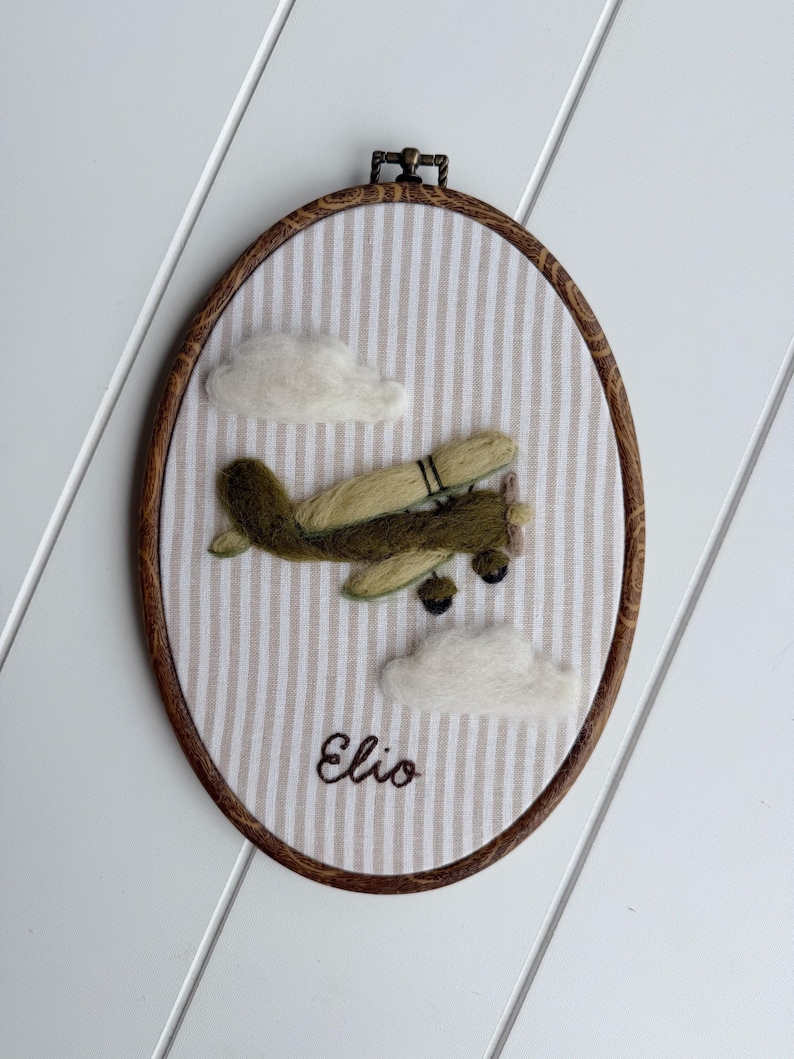 Puede incluir: Un bastidor de bordado ovalado con tela de rayas verticales beige y blancas. Un avi&oacute;n verde oliva de lana fieltrada vuela entre dos nubes de fieltro blanco. El nombre Elio est&aacute; bordado en cursiva marr&oacute;n oscuro en la parte inferior.