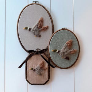 May include: Three framed wall hangings with felted duck designs. The ducks are brown, gray, and tan, with green and yellow accents. The frames are oval and rectangular, with brown wooden edges and neutral fabric backgrounds.