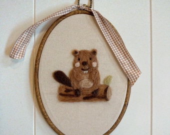 May include: An oval-shaped, framed artwork featuring a brown beaver sitting on a log. The beaver has a dark brown tail and a light brown body. The artwork is framed in a wooden hoop and has a gingham ribbon for hanging.
