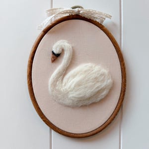 Op de afbeelding: Een wit zwanenkunstwerk van vilt, gemonteerd in een houten borduurring. De zwaan staat op een lichtroze achtergrond, met een bruine snavel en zwarte oogdetails. Bovenop is een crèmekleurige kanten strik bevestigd.