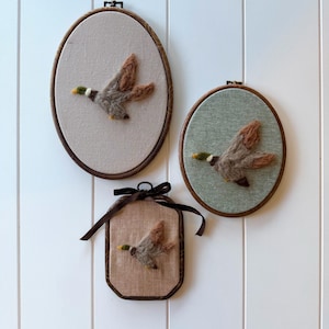 May include: Three framed wall hangings featuring felted ducks in flight. Two are oval, one is rectangular, all with brown wooden frames. The ducks have brown, gray, and yellow details, set against neutral fabric backgrounds. A brown ribbon adorns the rectangular frame.