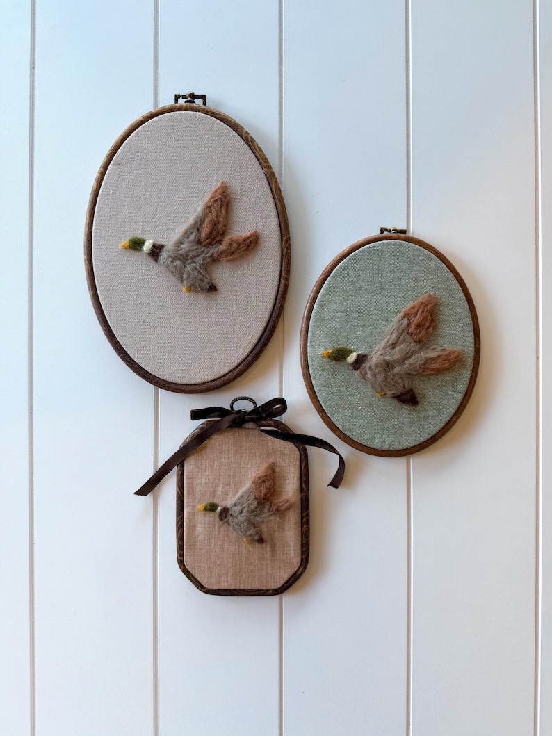 May include: Three framed wall hangings featuring felted ducks in flight. Two are oval, one is rectangular, all with brown wooden frames. The ducks have brown, gray, and yellow details, set against neutral fabric backgrounds. A brown ribbon adorns the rectangular frame.