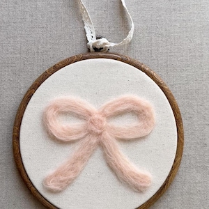 May include: A circular wooden embroidery hoop containing a piece of off-white fabric. Centred on the fabric is a soft, fuzzy, light pink bow made of fibre. A white lace ribbon is attached to the top of the hoop for hanging purposes.