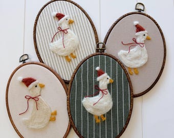 May include: Four oval-shaped wall hangings featuring felted white geese wearing red Santa hats and red ribbon bows. The geese have yellow feet and beaks. Each goose is set against a different fabric background, including striped and solid colors, within a wooden embroidery hoop.