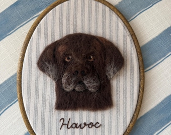 May include: An oval embroidery hoop frame containing a needle-felted portrait of a dark brown dog with a gray muzzle. The fabric background is white with thin blue vertical stripes. The name Havoc is embroidered in dark thread below the dog.
