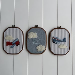 Puede incluir: Tres adornos de pared octogonales con marcos de madera oscura. Las piezas laterales muestran aviones y nubes de fieltro sobre tela de rayas azules y blancas. La pieza central tiene tela azul con nubes blancas y el texto Jack MONROE.