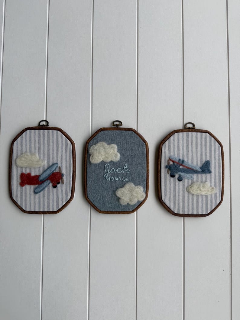 Puede incluir: Tres adornos de pared octogonales con marcos de madera oscura. Las piezas laterales muestran aviones y nubes de fieltro sobre tela de rayas azules y blancas. La pieza central tiene tela azul con nubes blancas y el texto Jack MONROE.