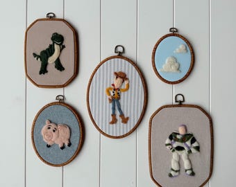 May include: Five needle felted wall hangings in wooden hoops featuring characters and motifs. The set includes a green dinosaur, a pink pig, a cowboy figure on striped fabric, a space ranger figure, and a light blue sky background with white clouds.