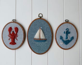 May include: Three oval embroidery hoops featuring nautical designs. The left hoop shows a red yarn lobster on blue and white striped fabric. The center hoop displays a felt sailboat on blue fabric. The right hoop features a blue yarn anchor.