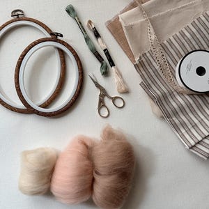 Könnte beinhalten: Zwei Stickrahmen aus Holz, zwei Stickgarnstränge in Grün und Creme, eine kleine verzierte Metallschere, eine Rolle weiße Spitzenborte, beigefarbene und gestreifte Stoffstücke sowie drei Wollvliesbüschel in Creme, Rosa und Hellbraun.
