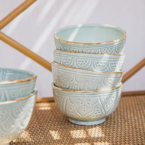 May include: A stack of six pale blue ceramic bowls with embossed floral patterns and gold rims. The bowls are stacked on a woven surface, with a bamboo structure in the background. The bowls are a light blue colour.