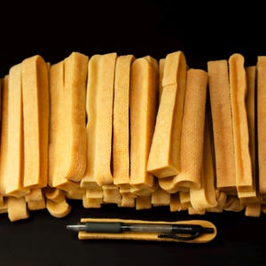 May include: A collection of light brown, rectangular chews, possibly dog treats, arranged in a stack against a black background. A black pen rests on a matching brown surface in the foreground. The chews have a textured appearance.