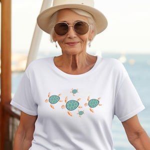 May include: White t-shirt featuring a design of several cartoon sea turtles in shades of teal and orange. The turtles are arranged in a horizontal pattern across the front of the shirt. The shirt is being worn.