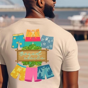 Pode incluir: Camiseta bege com um gráfico colorido de calções de banho e o texto "Bored Shorts". O gráfico apresenta vários pares de calções em azul, amarelo, rosa e verde, com estampas florais e de folhas. A camiseta é feita de um material macio.