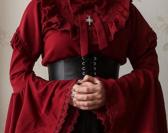 May include: A burgundy blouse with long, flared sleeves and lace trim. The blouse features a ruffled collar, a bow, and a cross pendant. A black corset belt cinches the waist. The outfit is completed with black bottoms.