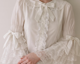 May include: A white blouse with a Peter Pan collar, adorned with a satin bow at the neckline. The long sleeves feature lace trim and bows at the cuffs. The front of the blouse has a row of buttons and lace detailing.