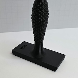 Puede incluir: Un sello negro con un mango texturizado y una base rectangular. El mango tiene un patrón de diamantes. La base tiene el texto "M55" impreso. El sello está hecho de un material oscuro y sólido.