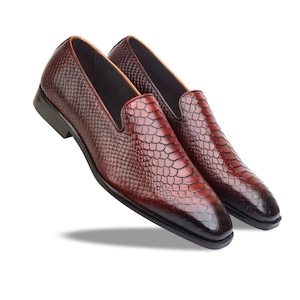 May include: A pair of dark burgundy loafers featuring a textured snakeskin pattern across the upper surface. The shoes have a pointed toe, a low heel, and a smooth dark interior lining, showcasing a polished finish with a gradient colour effect.
