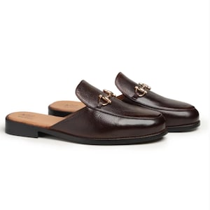 May include: A pair of dark brown leather backless loafers featuring a textured pebble grain finish and a gold-toned metal horsebit detail across the vamp. The shoes have a low heel, a rounded toe, and a tan interior lining with visible branding.