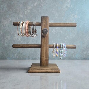 May include: A dark brown wooden bracelet stand with two levels for displaying jewellery. The stand holds a selection of bracelets in shades of blue, pink, white, and purple. The background is a muted blue-grey.