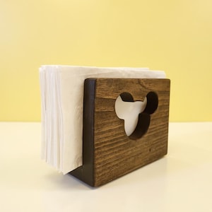 以下が含まれることがあります： 濃い茶色の木製ナプキンホルダー。マウスの頭の形に切り抜かれています。ホルダーの中には白いナプキンの束があります。背景は薄い黄色です。木製のホルダーです。
