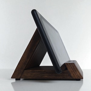 May include: A dark brown wooden tablet stand in a triangular shape. The stand is designed to hold a tablet at an angle for easy viewing. The tablet is black and rests securely in the stand. The stand is made of wood and has a smooth finish.