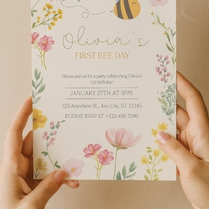 Può includere: Un invito floreale con il testo "Olivia's FIRST BEE DAY". Il design include un'ape dei cartoni animati, elementi floreali e i dettagli dell'evento. La carta è tenuta da due mani.