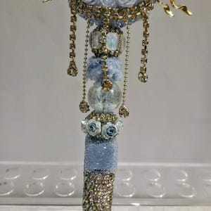 May include: A decorative pen with a gold and rhinestone butterfly design. The pen features a light blue and gold colour scheme, with rhinestones and faux pearls. The pen is embellished with small blue roses and dangling rhinestone chains.