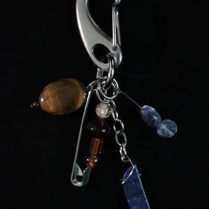 May include: A silver-toned metal carabiner keychain featuring a collection of dangling charms. The charms include a smooth brown tiger's eye bead, a safety pin with stacked dark red and clear beads, a small silver chain, and a blue stone pendant.