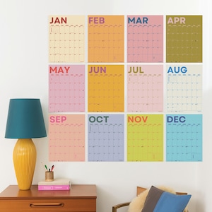 May include: A set of twelve monthly calendars, each with a different pastel background colour and the month's name in bold letters. The calendars are displayed on a white wall, with a yellow lamp and a wooden table in the foreground.