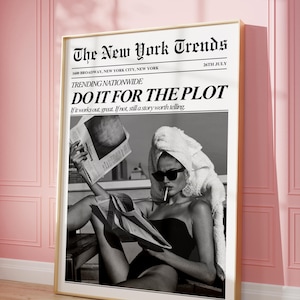 May include: A framed black and white print featuring a woman in sunglasses and a towel reading a newspaper. The print has the text "The New York Trends" and "DO IT FOR THE PLOT". The frame is gold and the background is a pink wall.