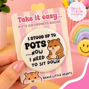 May include: A round pinback button featuring a cartoon hamster and the text I STOOD UP TO POTS....NOW I NEED TO SIT DOWN. The button is attached to a pink and white striped backing card labeled Take it easy... POTS AWARENESS BADGE.
