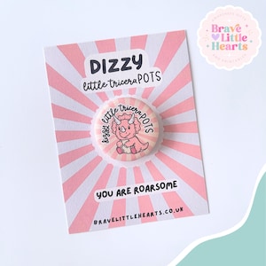 May include: A pink and white button with a cartoon triceratops illustration, text that reads "Dizzy little tricera POTS", and the phrase "You are roarsome". The button is on a pink and white striped card.