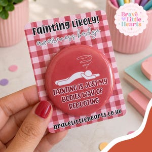 May include: A round red pin-back button on a red and white gingham display card. The button features a white graphic of a person lying down with a swirl above their head and the text: FAINTING IS JUST MY BODIES WAY OF REBOOTING.